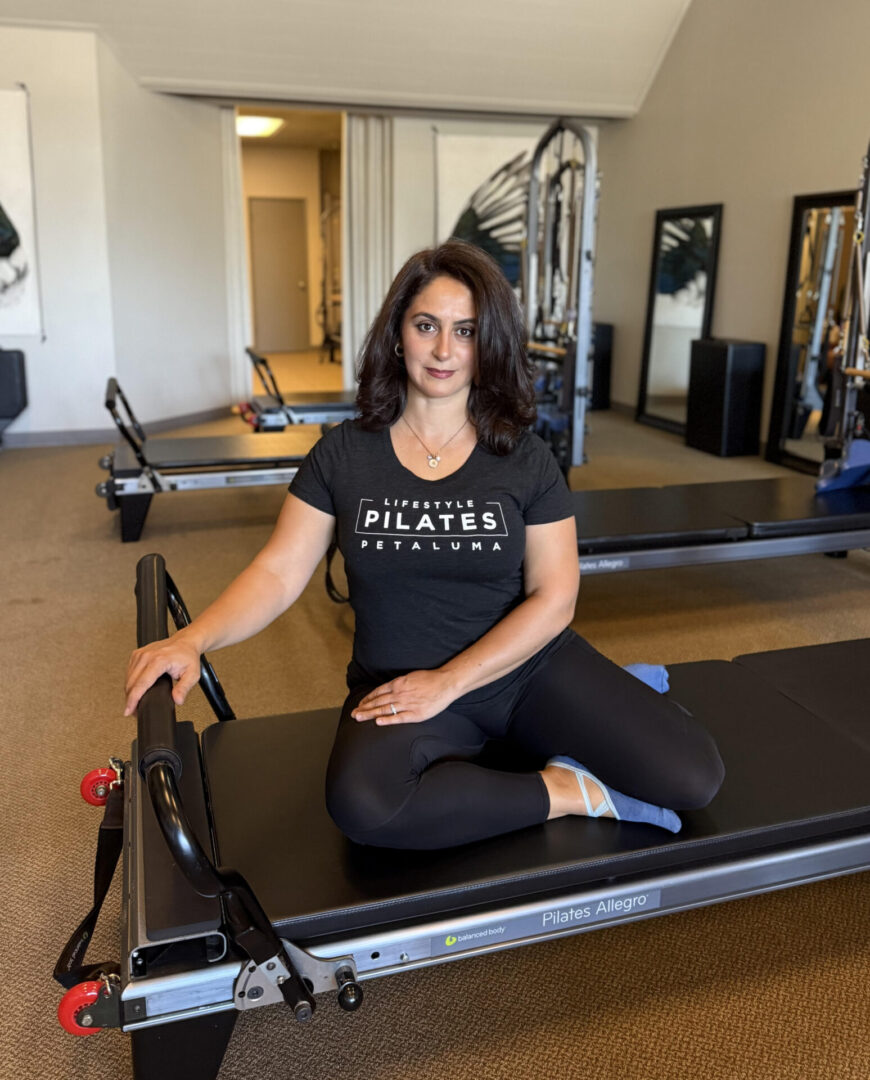 Woman in black Pilates attire sitting on a reformer in a studio.
